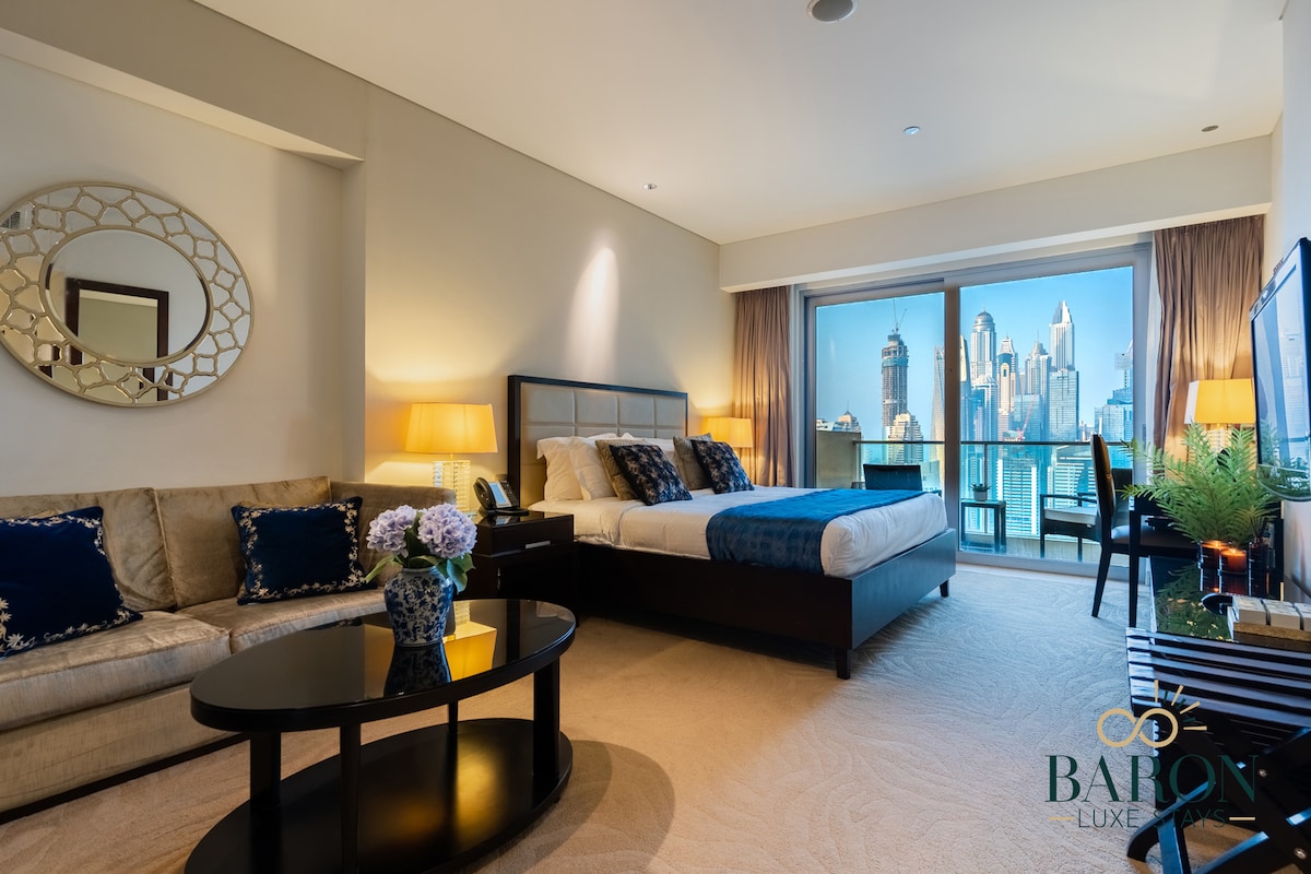 A spacious studio apartment is featured, showcasing a king-size bed with plush linens and decorative pillows. A comfortable seating area with a beige sofa and a round coffee table is visible, along with large windows offering stunning views of the city skyline.
