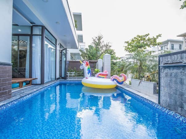 A clear blue infinity pool is featured, with a floating inflatable unicorn adding a playful touch. The pool area is surrounded by greenery and spacious decks, with large glass doors providing access to the indoors and enhancing the connection to the outdoor space.