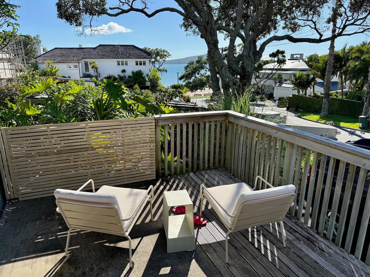 An outdoor deck features two light-colored chairs positioned around a small table. Lush greenery frames the area, with views of a beach and sparkling waters visible in the background under a clear blue sky.
