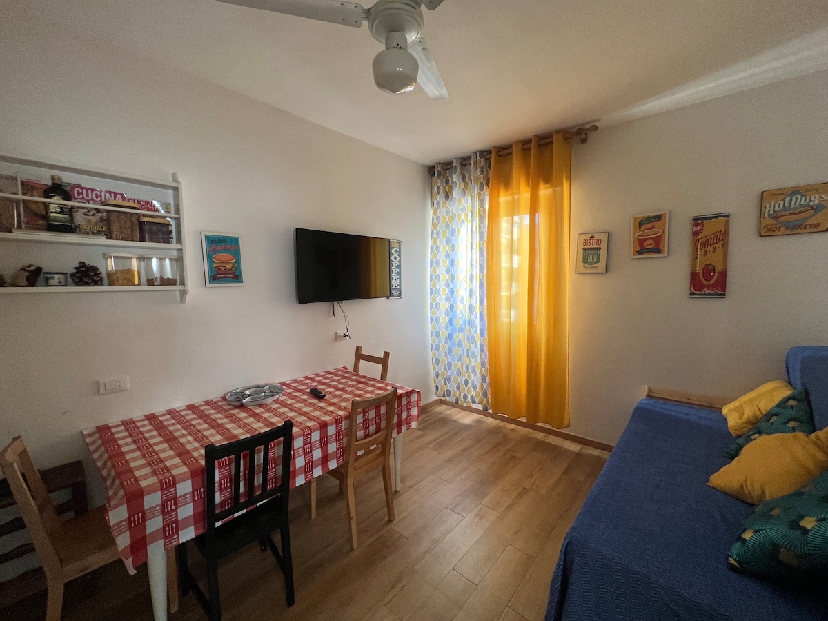 A dining area with a checkered tablecloth is complemented by a flat-screen TV mounted on the wall. Bright yellow curtains frame the window, adding color to the light-filled room. A comfortable seating area is shown, decorated with colorful cushions.