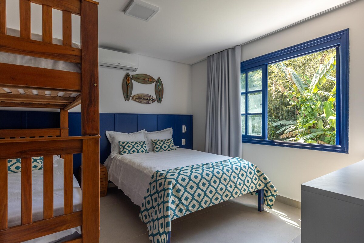 A well-lit suite features a spacious bed and a bunk bed against a blue accent wall. Natural light enters through a large window, revealing lush greenery outside. Decor includes fishing-themed wall art, with neutral-toned bedding and a geometric-patterned throw complementing the serene atmosphere.