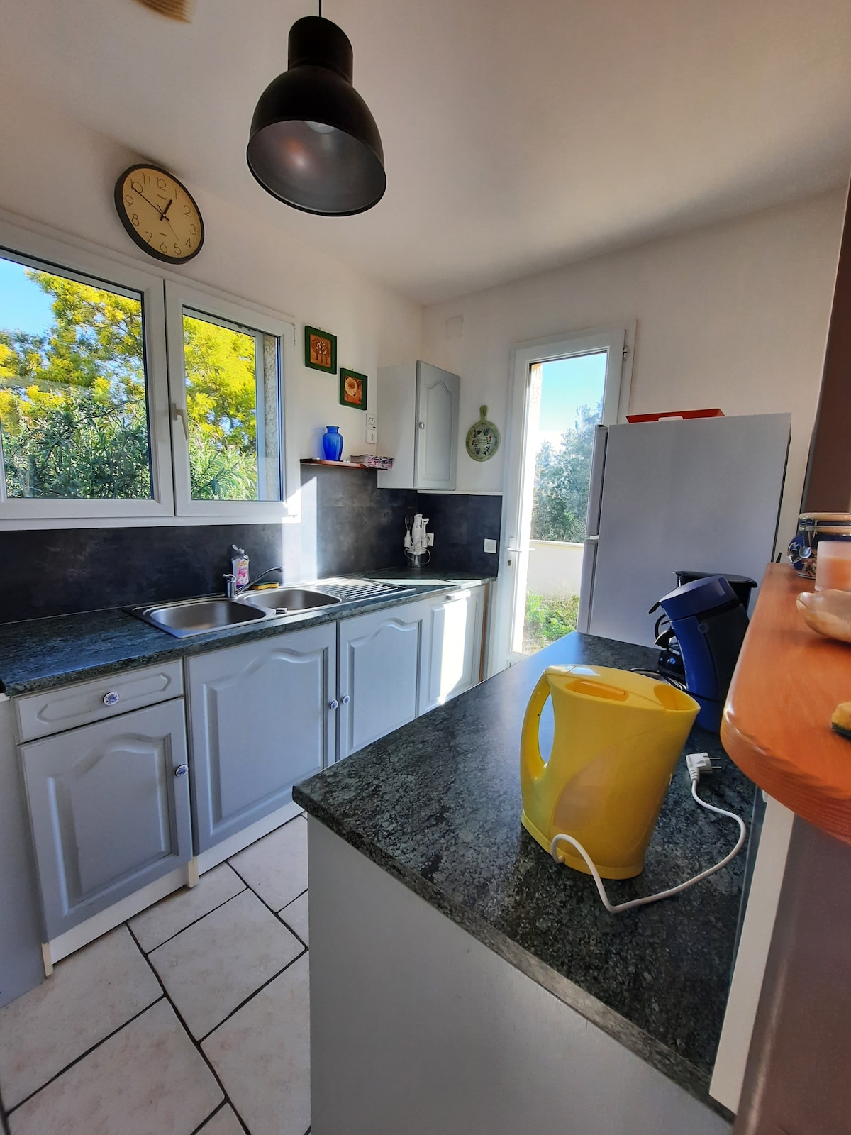 A well-equipped kitchen features light-colored cabinetry and a dark countertop. Natural light enters through multiple windows, illuminating the space. A coffee maker and a yellow kettle are placed on the countertop, while modern appliances, including a fridge, are visible in the background.