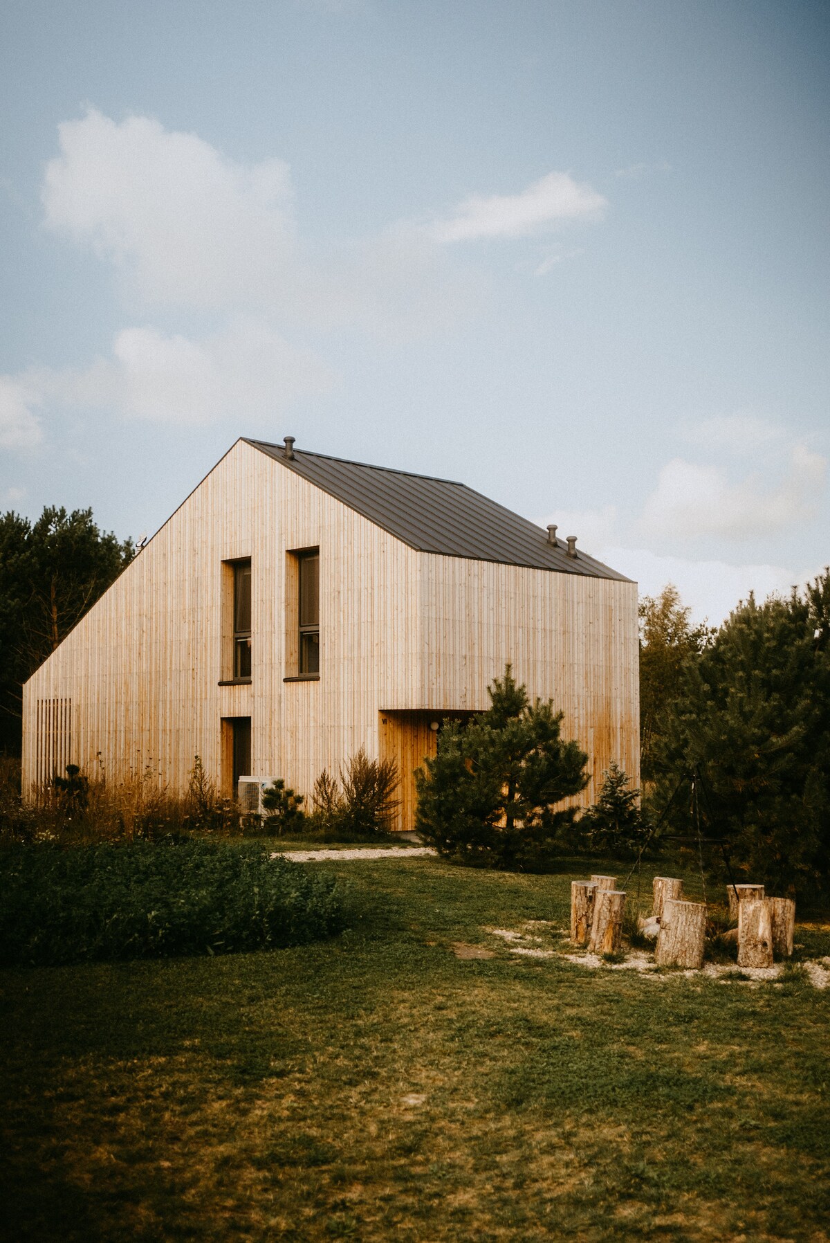 A modern wooden house features a unique triangular roof, complemented by large windows. Surrounding greenery includes tall trees and shrubs, contributing to the serene setting. A small clearing with logs is visible in the foreground, suggesting areas for seating or gathering.