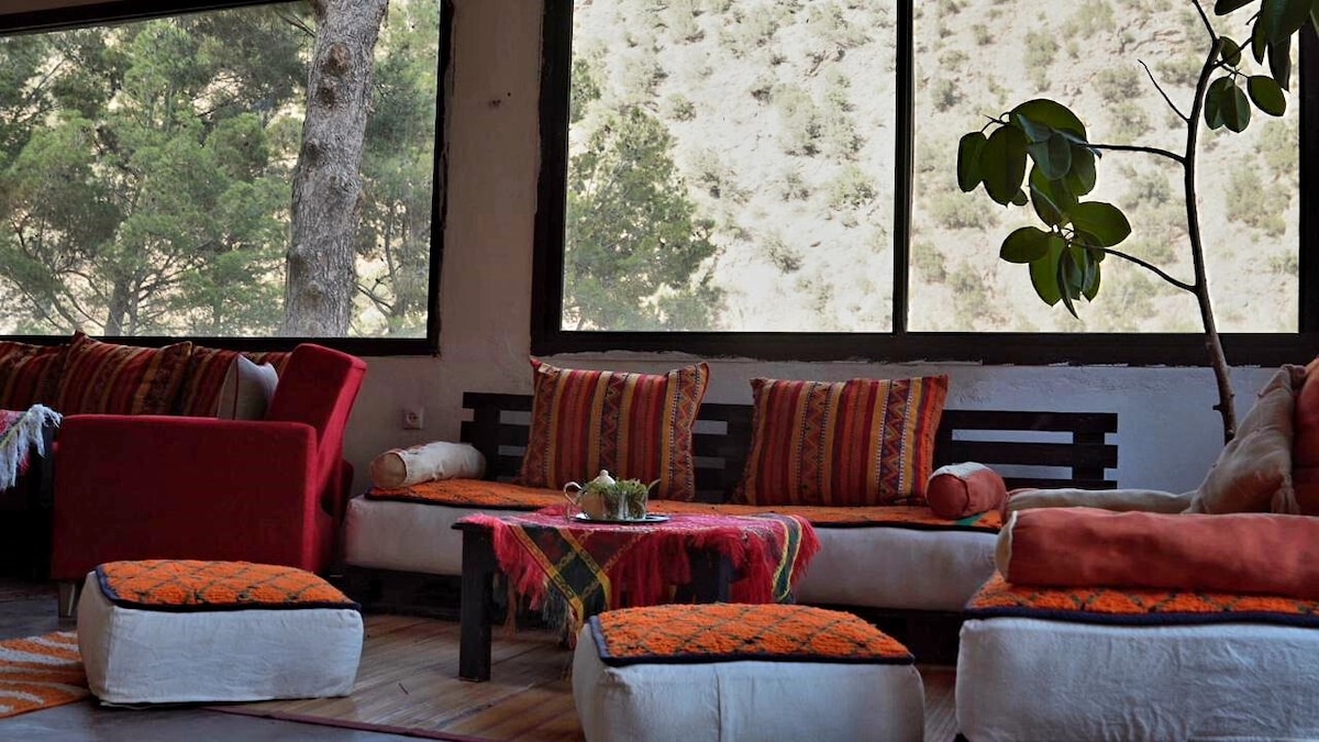 A cozy living area features large windows offering views of the surrounding landscape. Colorful cushions adorn low seating areas, while a small coffee table is set in the center. Warm, earthy tones are complemented by natural light filtering through the glass.