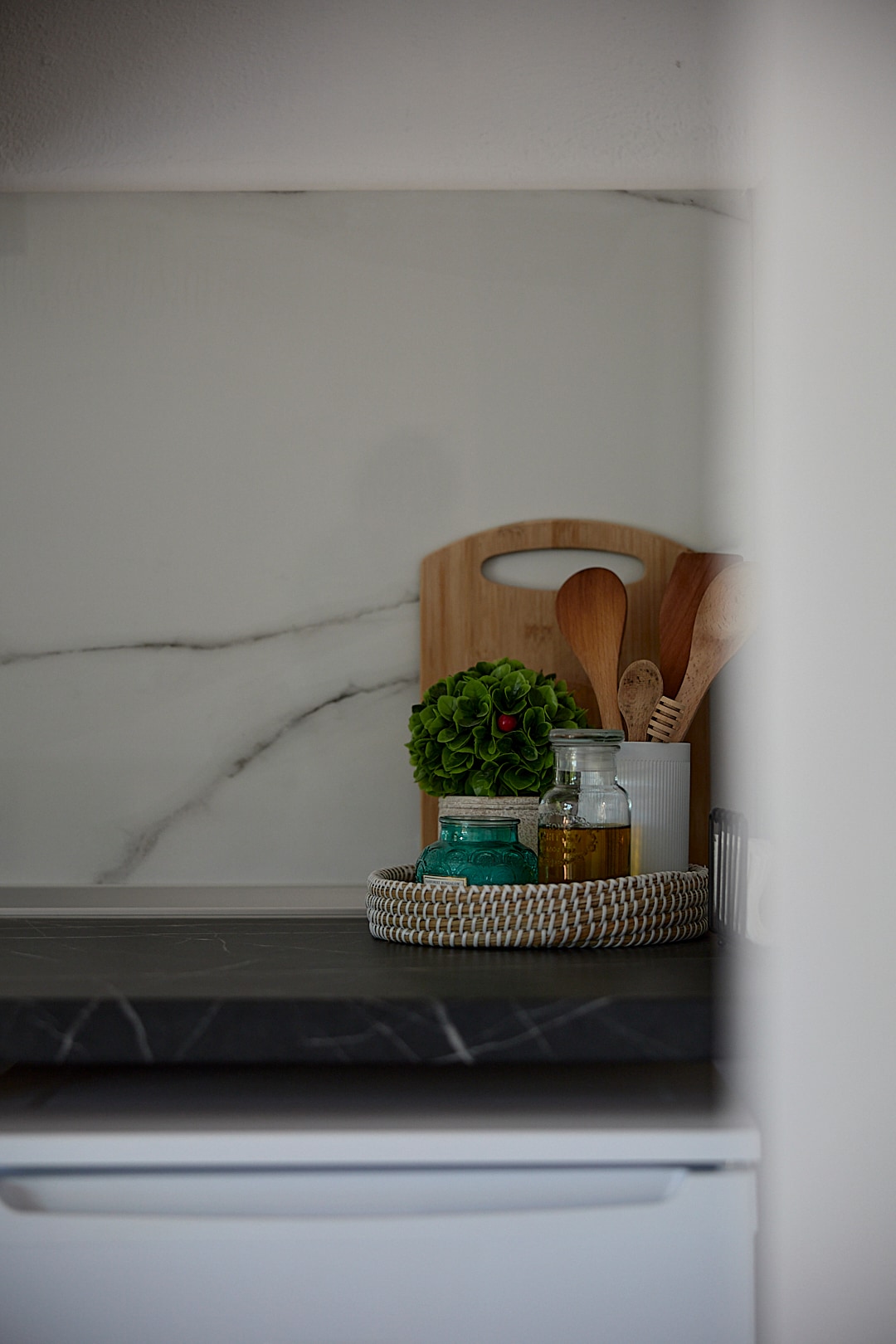 A kitchen area is showcased featuring dark countertops and a subtle marble backsplash. A wooden cutting board and utensils are arranged alongside decorative items, including a green plant, in a woven basket. Various jars and bottles are included for cooking and storage.