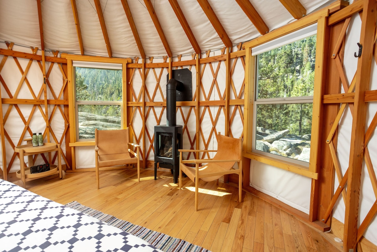 Inside the yurt, cozy seating areas are arranged with two wooden chairs positioned beside a gas fireplace. Natural light floods through two large windows, highlighting the wooden flooring and the yurt's unique architecture, featuring a warm, earthy color palette.