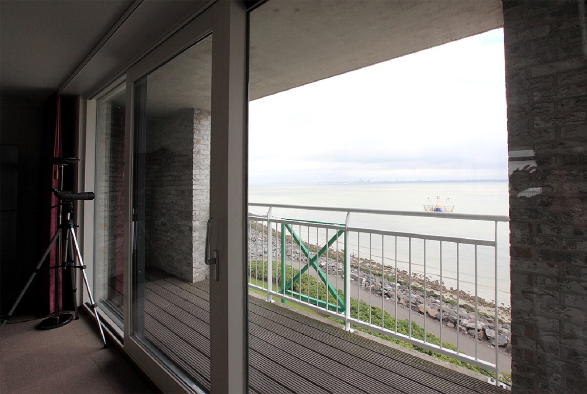 A large glass door opens to a private balcony overlooking the harbor. A telescope is positioned nearby to enhance views of passing ships and, on clear days, the coast of Vlissingen. The calm water and distant landscape are visible beyond the railing.