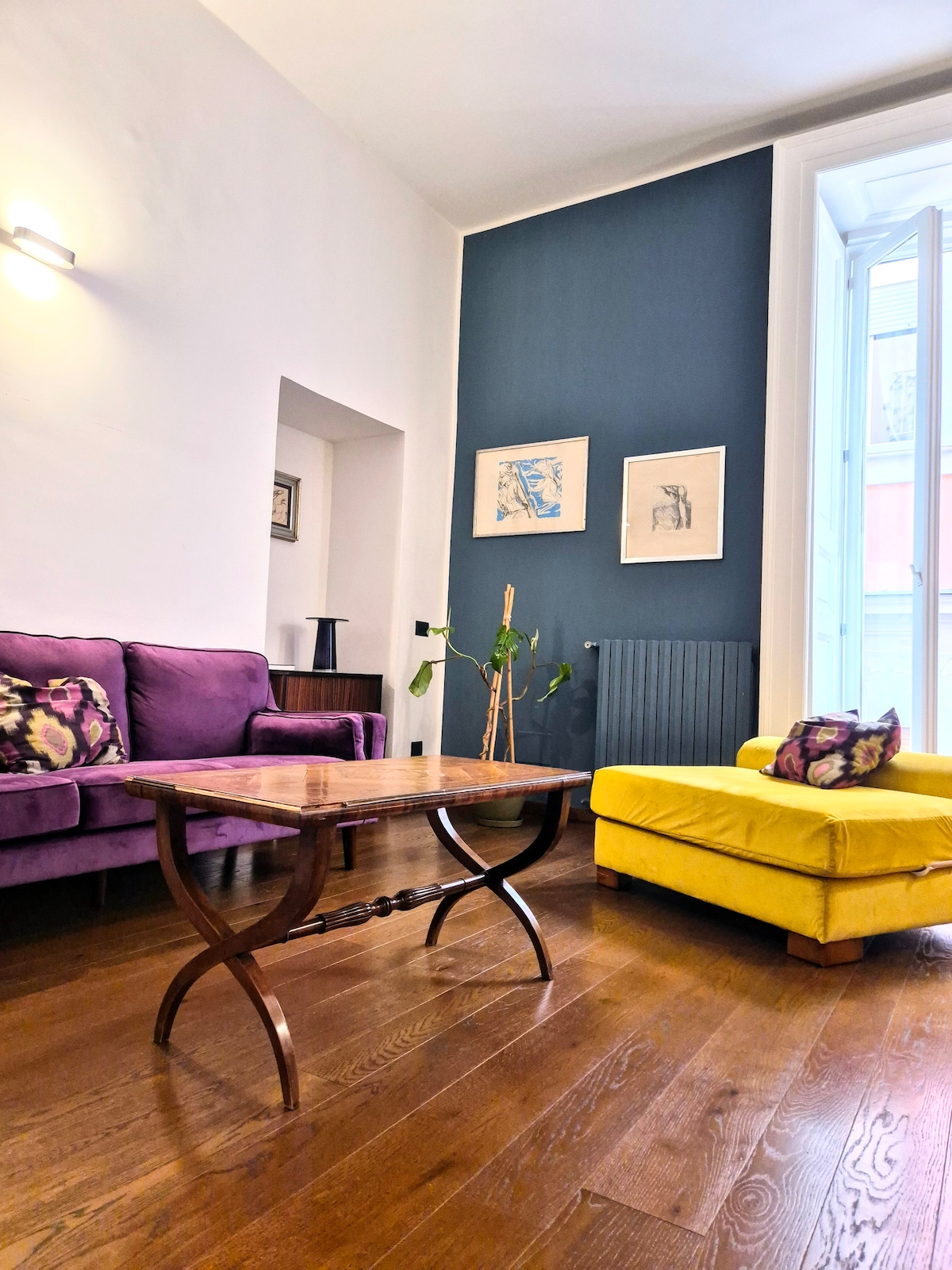 The living area features a mix of seating options, including a purple sofa and a yellow armchair. A wooden coffee table sits in the center. Light streams through the open window, illuminating the space which is accented by dark blue and white walls.