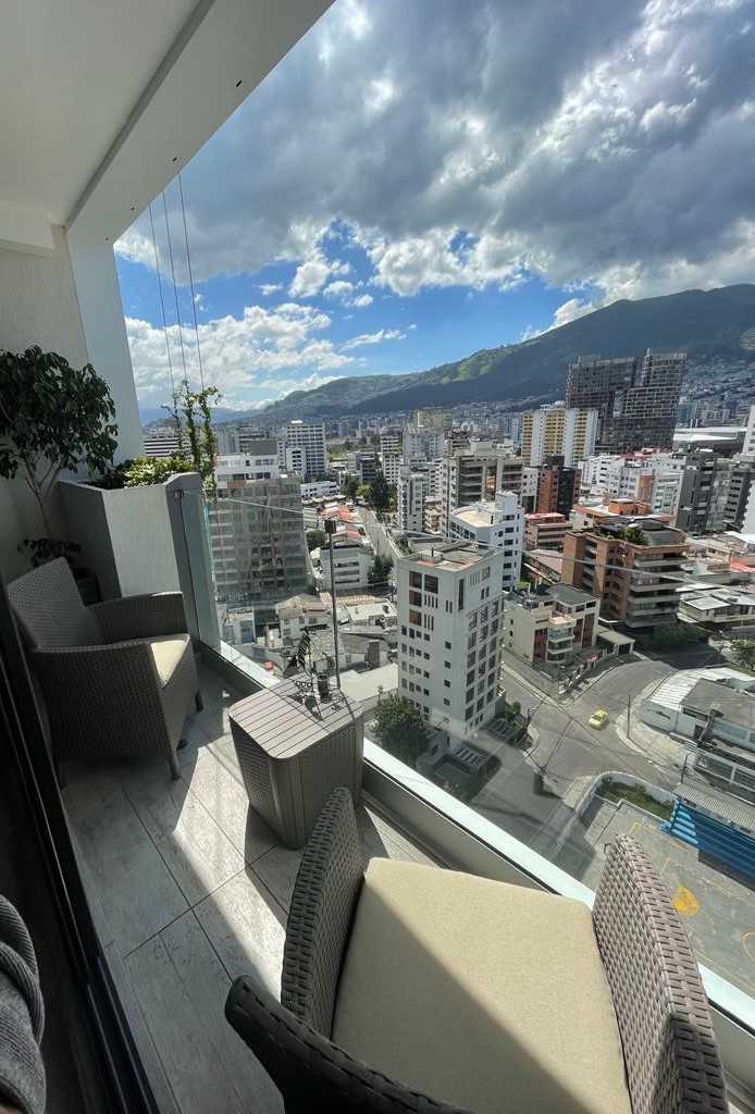 A balcony area is furnished with two wicker chairs and a small table, offering panoramic views of the city and surrounding mountains. The glass railing provides safety while allowing unobstructed visibility. Natural light brightens the space, with clouds adding texture to the sky.