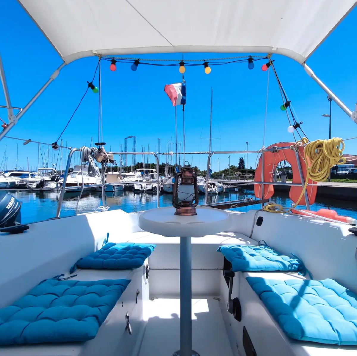 The cockpit of the sailboat features blue cushioned seating around a circular table, creating a comfortable gathering space. Brightly colored lights are strung overhead, and a traditional lantern is placed on the table. Boats can be seen docked in the marina under a clear blue sky.