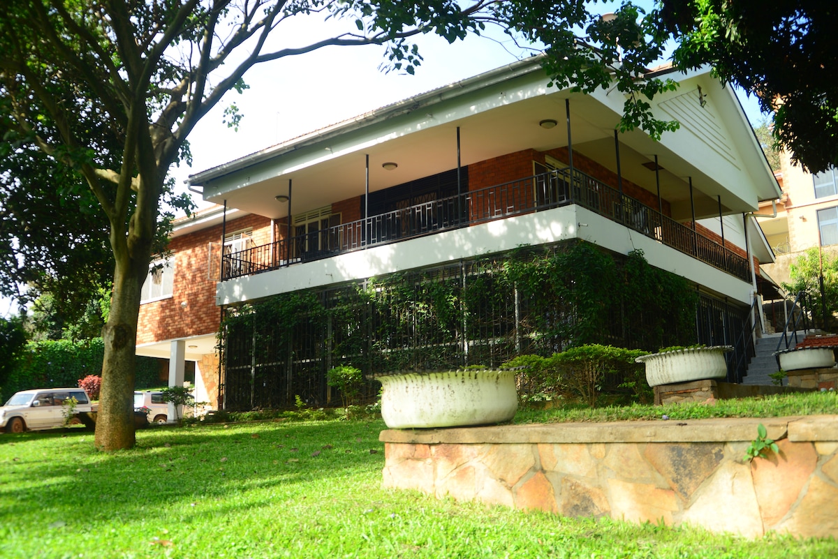 A spacious two-story villa is shown, featuring a large balcony with black wrought iron railings. Lush green lawn surrounds the property, with mature trees providing shade. The facade displays a combination of brick and white siding. A stone seating area is visible in the foreground.