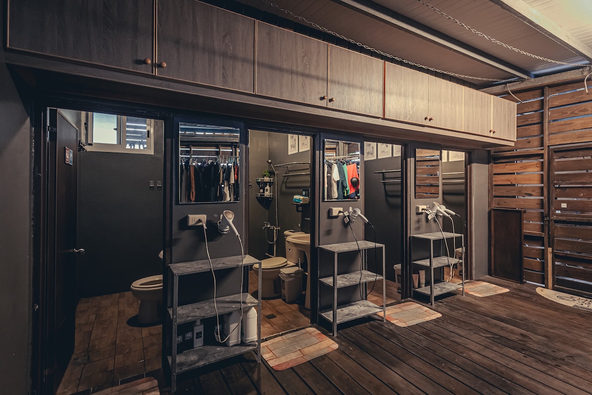 A shared bathroom space showcases three individual stations, each equipped with a sink and a mirror. Shelving units provide storage for personal items, while a toilet is visible in the background. The wooden flooring complements the industrial design, creating a functional and organized environment.