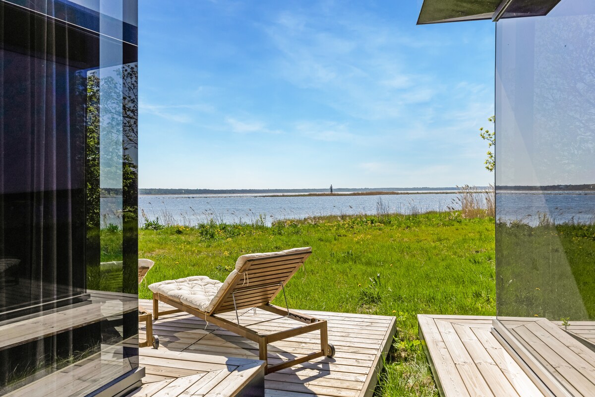 A sunny outdoor area features a wooden lounge chair positioned on a deck overlooking a grassy expanse leading to a serene water view. Clear glass walls provide a seamless connection to the landscape, framing the tranquil scenery beyond.