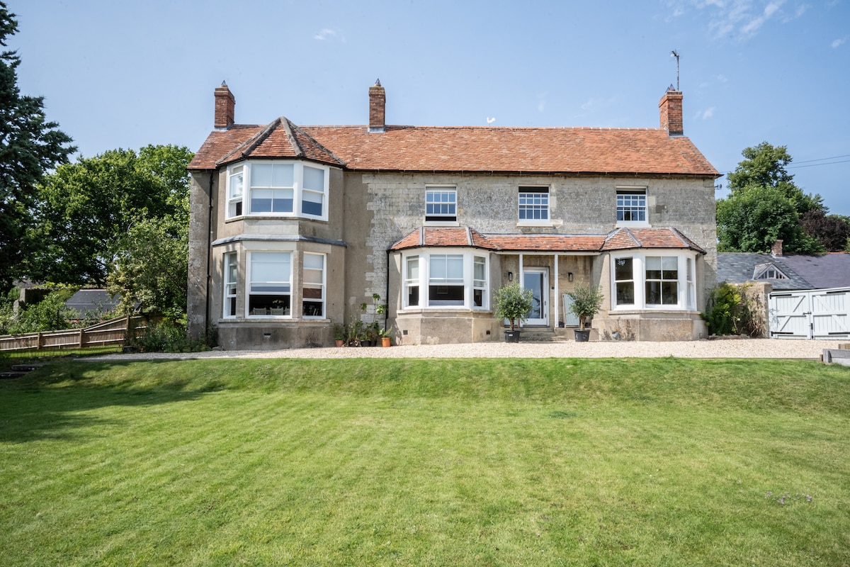 A charming, two-story rural home is set against a backdrop of lush green grass. The façade features multiple large windows and a classic roof, providing a welcoming appearance. Surrounding trees and landscaped gardens enhance the outdoor space.