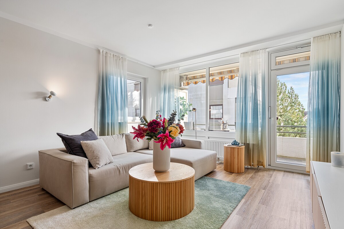 A modern living area is highlighted by a light-colored sofa adorned with accent pillows. A circular wooden coffee table is centered on a soft area rug, while large glass doors open to a balcony with a view. Natural light fills the space, illuminating the soft gradient curtains.