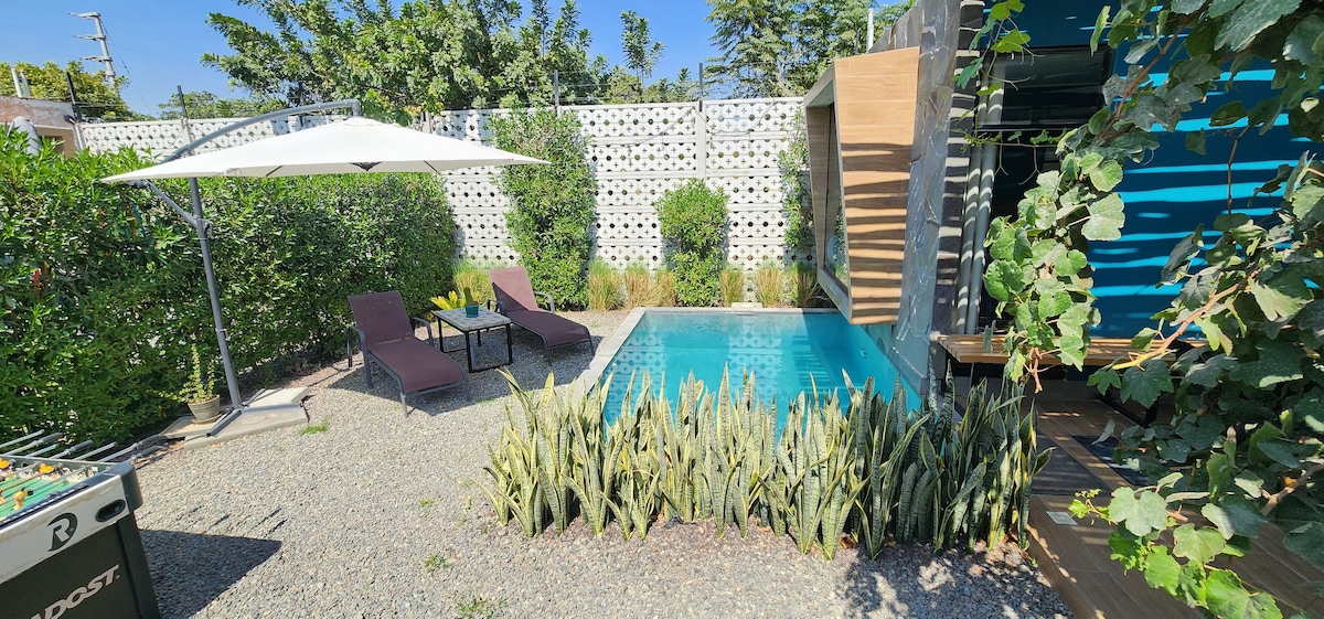 An outdoor area is presented, featuring a private pool surrounded by ornamental plants and gravel. Two lounge chairs are positioned beside the pool, complemented by a shade umbrella. The space is enclosed by greenery, providing privacy in a serene setting.