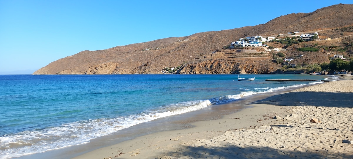 A sandy beach is depicted, with gentle waves lapping at the shore. The clear blue water contrasts with the tan sand. Rolling hills provide a backdrop, dotted with white buildings. A small boat is visible in the water, contributing to the tranquil coastal scene.