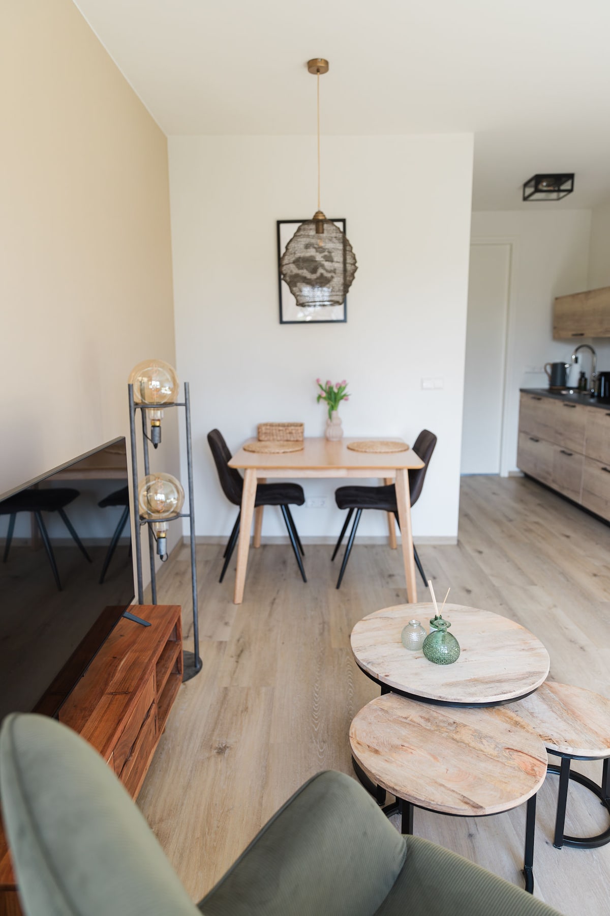 A dining area features a wooden table surrounded by four black chairs. A vase with fresh flowers decorates the table. Two rustic wood side tables are placed nearby, and a minimalist light fixture hangs from the ceiling, enhancing the modern aesthetic.