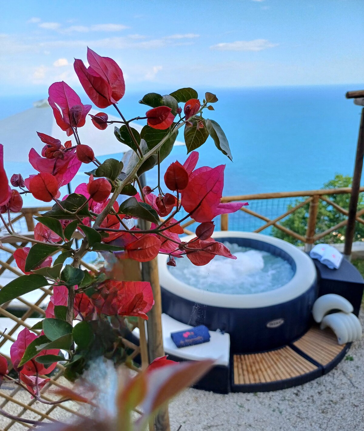 A private jacuzzi is situated on a wooden deck, surrounded by vibrant bougainvillea flowers. The tranquil water is bubbling, and the view extends over the blue sea, providing a serene atmosphere for relaxation.