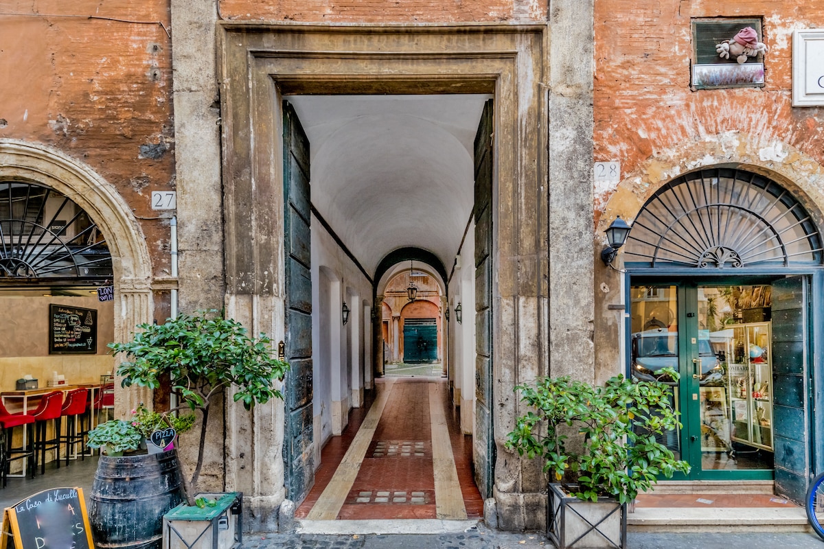 An entrance to a historic building is framed by stone walls, showcasing an arched corridor. The floor features a patterned design, leading towards an illuminated passageway. Potted greenery stands on either side, enhancing the inviting entry. Wooden doors are visible at the far end.