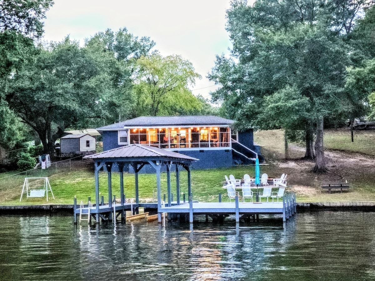 The cozy lake home is presented in a tranquil setting, surrounded by trees. A spacious dock extends into the water, featuring seating and an umbrella. The home, with large windows, showcases a welcoming atmosphere as evening light glows from within.