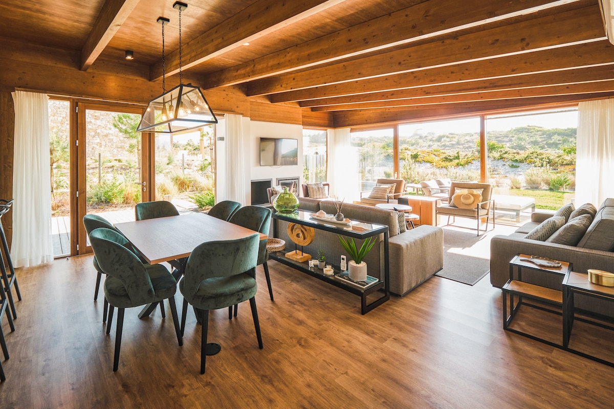 An open-concept living area features a large dining table surrounded by upholstered chairs. Natural light floods the space through expansive glass doors, revealing views of the outdoor landscape. A comfortable seating arrangement is positioned to invite relaxation, complemented by natural wood beams overhead.