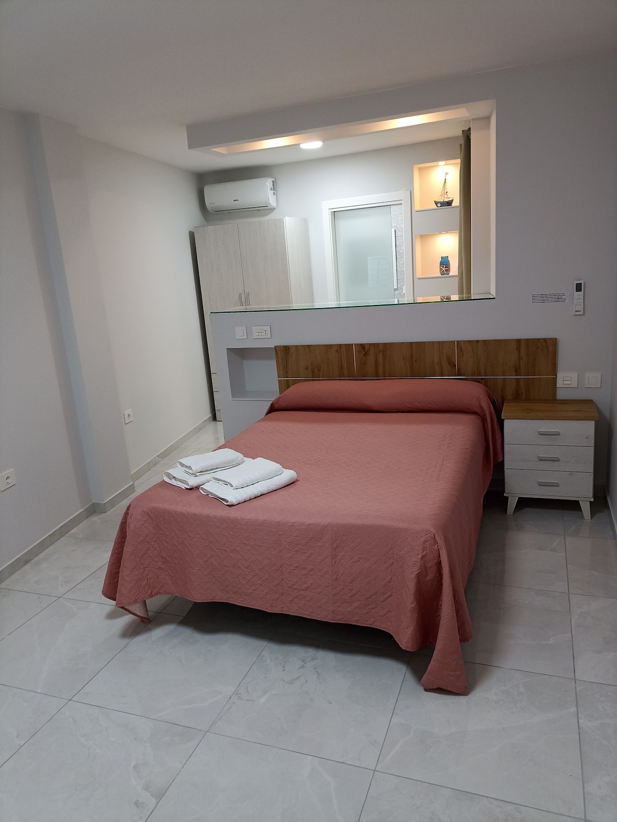 A cozy sleeping area features a spacious bed dressed in soft coral linens. Fresh towels are neatly folded on the bed. Light-colored furniture complements the soothing palette, while a wall-mounted air conditioning unit provides comfort. Natural light illuminates the room through a nearby window.