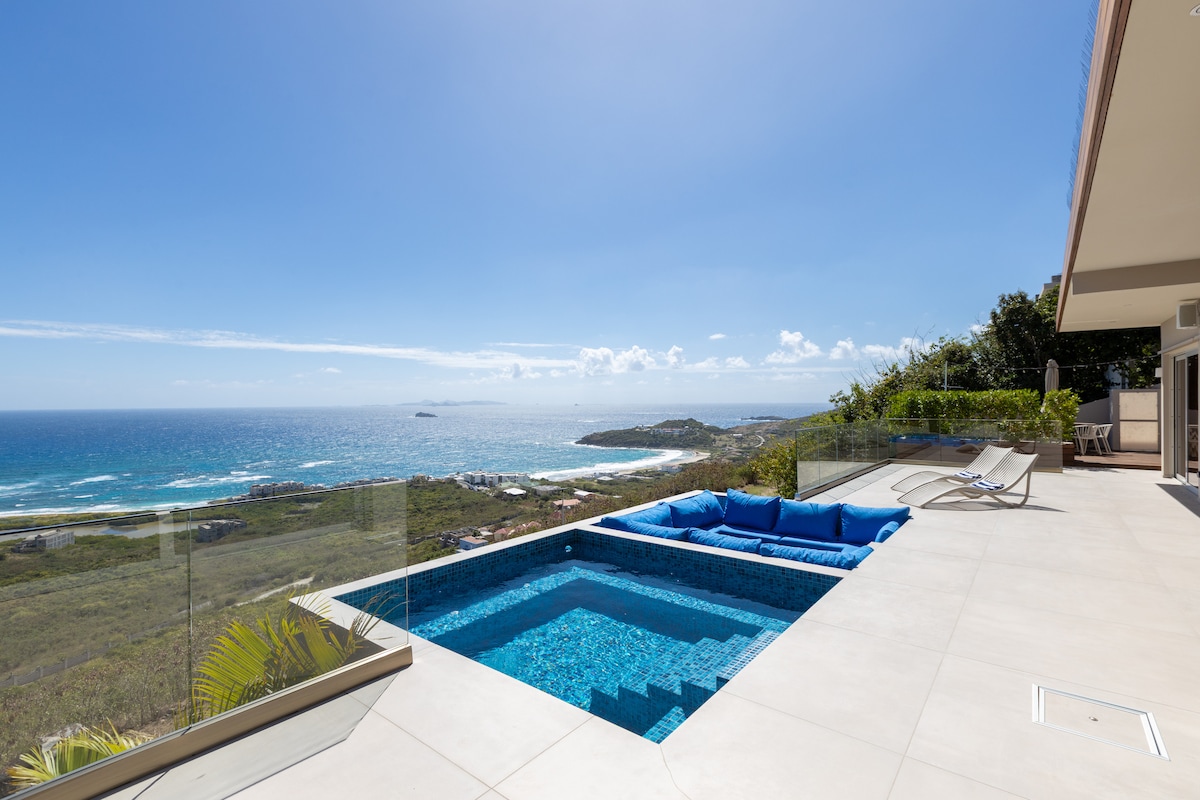 An outdoor terrace features a private, heated plunge pool surrounded by sleek stone tiles. Comfortable blue seating borders the pool, with a clear glass railing offering unobstructed views of the ocean and coastline under a bright blue sky.