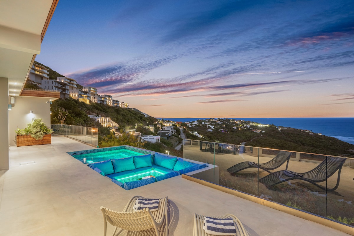 An outdoor terrace features a serene pool set against a backdrop of rolling hills and the ocean. Comfortable lounge chairs are positioned nearby, creating an inviting space for relaxation. Colorful sunset hues paint the sky, enhancing the tranquil atmosphere.
