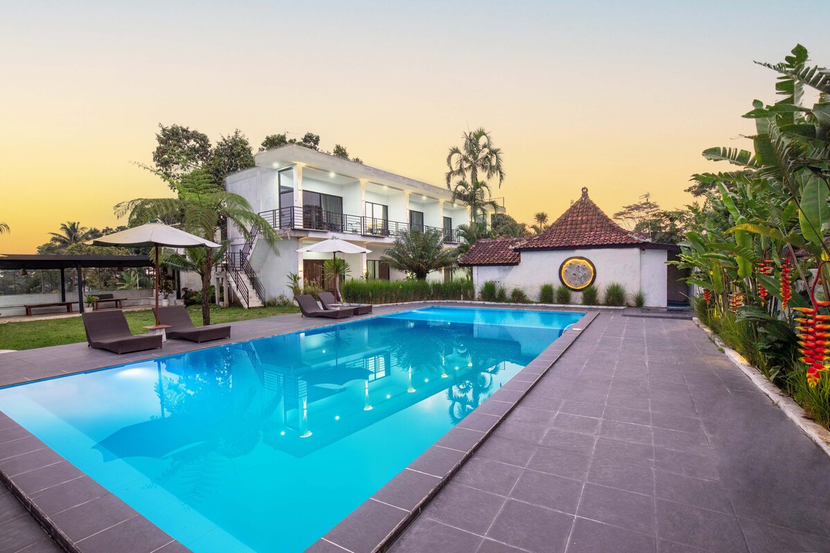 A refreshing swimming pool is surrounded by lush greenery and lounge chairs, providing relaxation opportunities. In the background, a modern two-story building is positioned alongside a traditional structure with a distinctive roof, framed by palm trees and a serene evening sky.