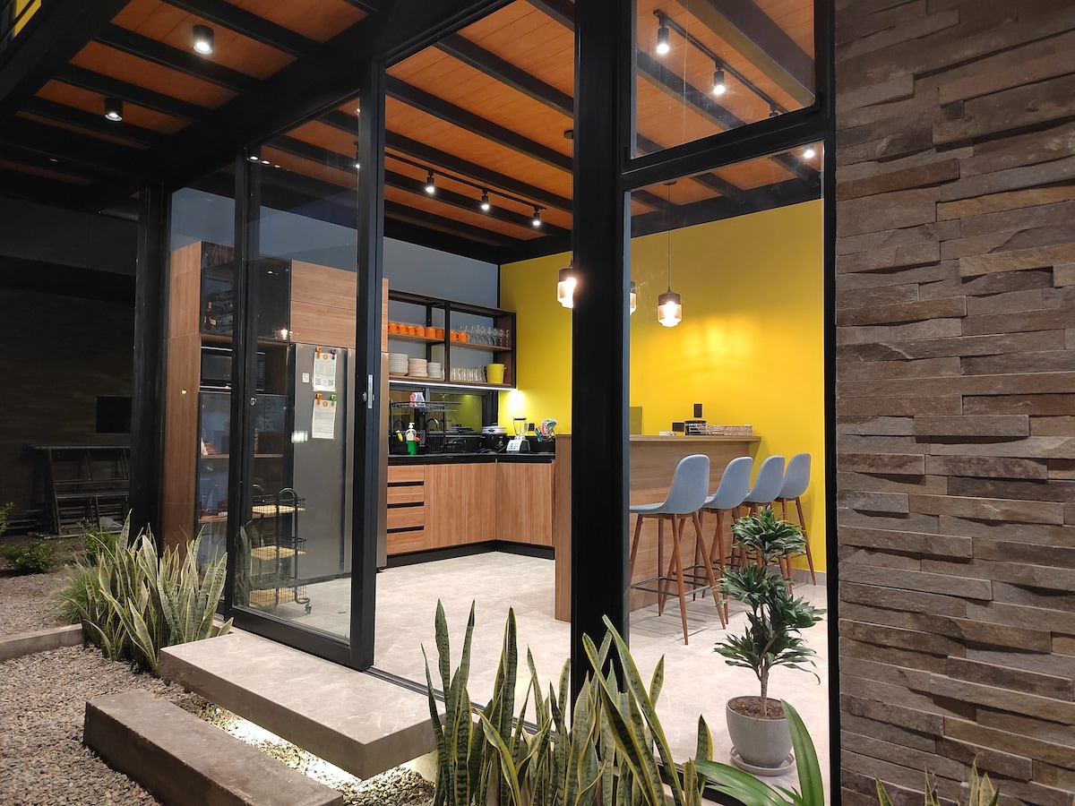 A modern kitchen is featured with wooden cabinetry and a spacious counter area. Three blue bar stools are situated at a sleek counter. Bright yellow walls add a vibrant touch, while large glass doors create an open connection to the outdoor space.