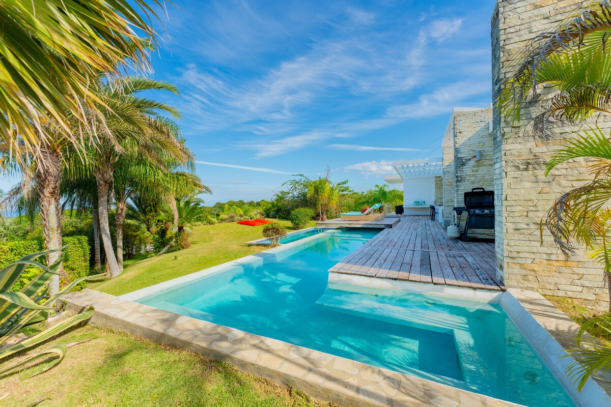An inviting infinity-style pool reflects the clear blue sky, surrounded by lush landscaping. A spacious wooden deck extends along one side, providing areas for seating and relaxation. Tropical plants frame the scene, enhancing the serene atmosphere of this outdoor space.