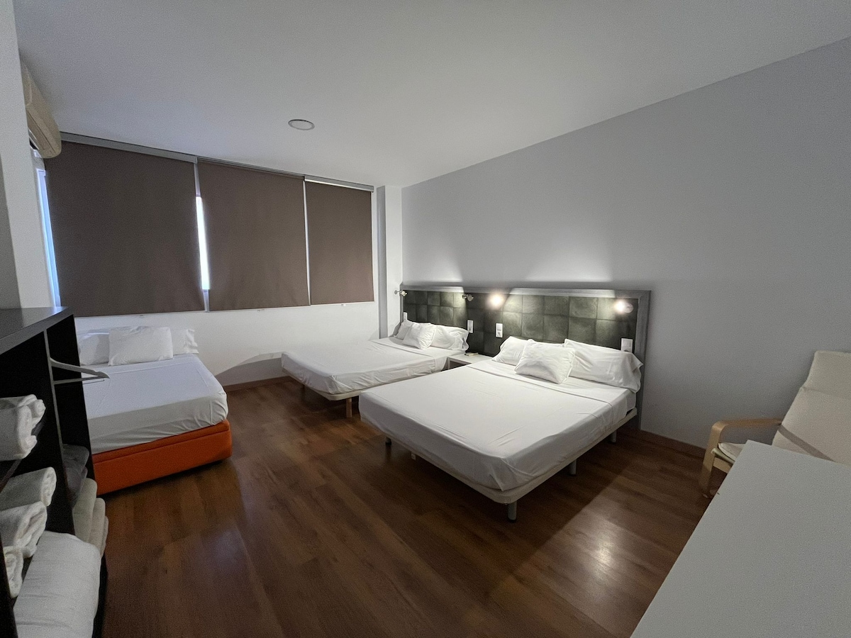The room features multiple beds: two spacious beds, each with gray upholstered headboards, and a single bed positioned alongside. Soft natural light enters through large windows fitted with brown shades. A minimalist white chair and a black shelving unit are visible in the corner.