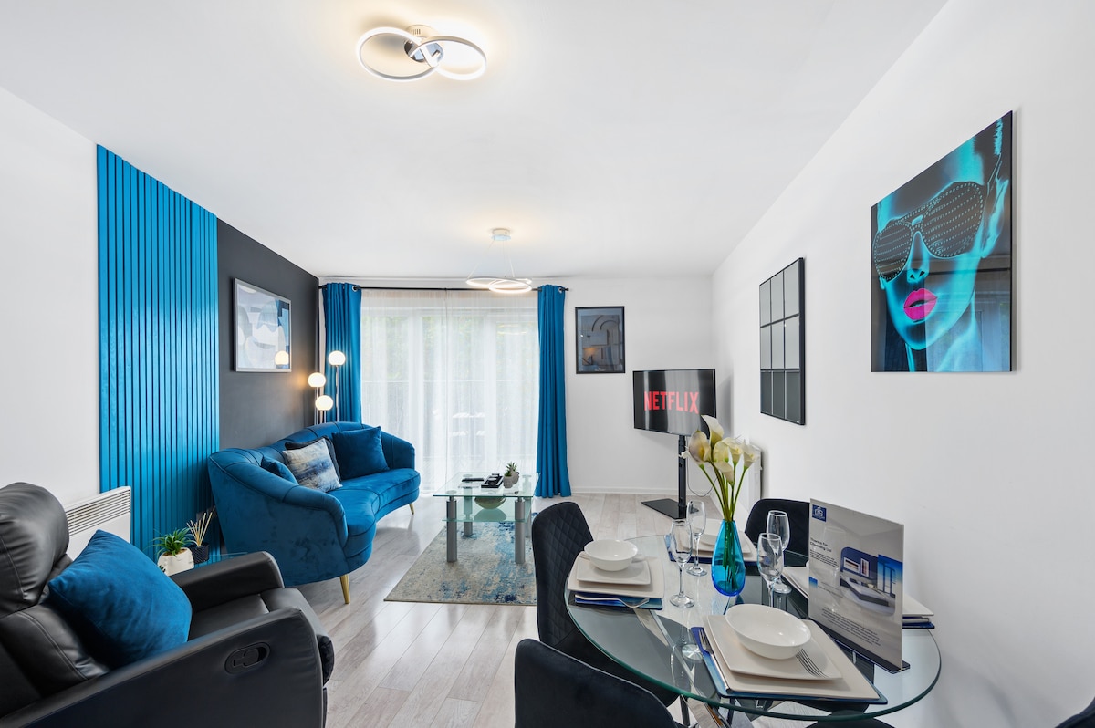 A modern living area features a blue sofa and a glass coffee table, set against a backdrop of blue and black walls. Natural light enters through large windows, with a Netflix-enabled TV visible. A dining table is elegantly set for four, enhancing the inviting atmosphere.