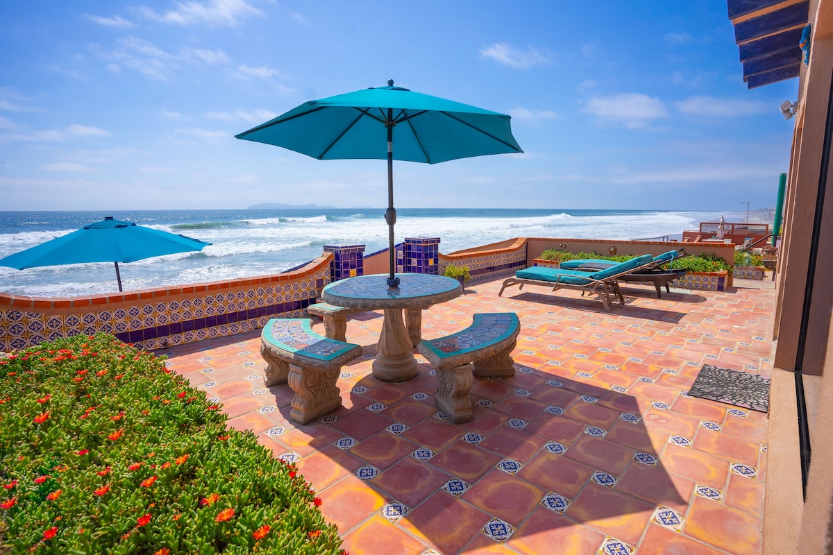 A sunlit outdoor patio features a stone dining table surrounded by colorful tile work. Two vibrant umbrellas provide shade for seating areas, and lounge chairs invite relaxation. Lush greenery adds a natural touch, with the waves of the ocean visible in the background.