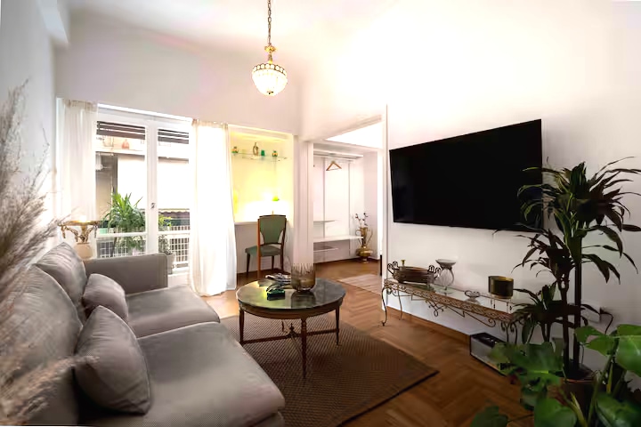 A spacious living area features a comfortable, light-colored sofa positioned near a stylish round coffee table. A large TV is mounted on the wall, and ample natural light enters through glass balcony doors. Greenery and decorative elements add warmth to the space.