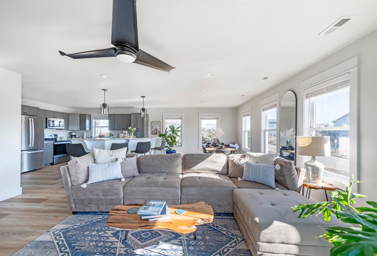 A modern living area features a large sectional sofa arranged around a natural wood coffee table. Light-colored walls and hardwood flooring complement the sleek kitchen in the background. Large windows allow natural light to fill the space, creating an inviting atmosphere.