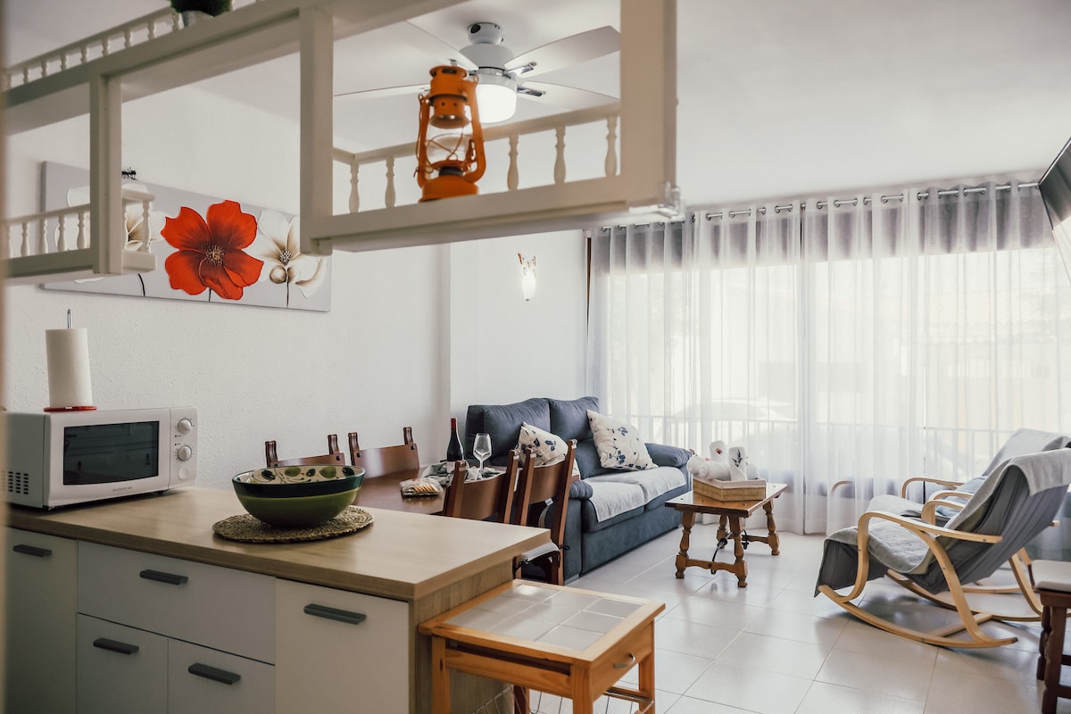 An open-plan living area is presented, featuring a comfortable sofa and rocking chair situated by large windows covered with sheer curtains. A wooden table is placed next to a microwave, while a decorative bowl sits at the counter. A warm atmosphere is created by floral wall art.