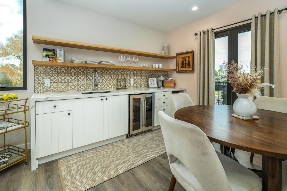 A modern kitchen area features light cabinetry with an open shelf displaying glassware and decor. A wine refrigerator is positioned next to a sink, complemented by patterned backsplash tiles. A round dining table with plush chairs sits across from the kitchen, adorned with a simple vase of dried flowers.