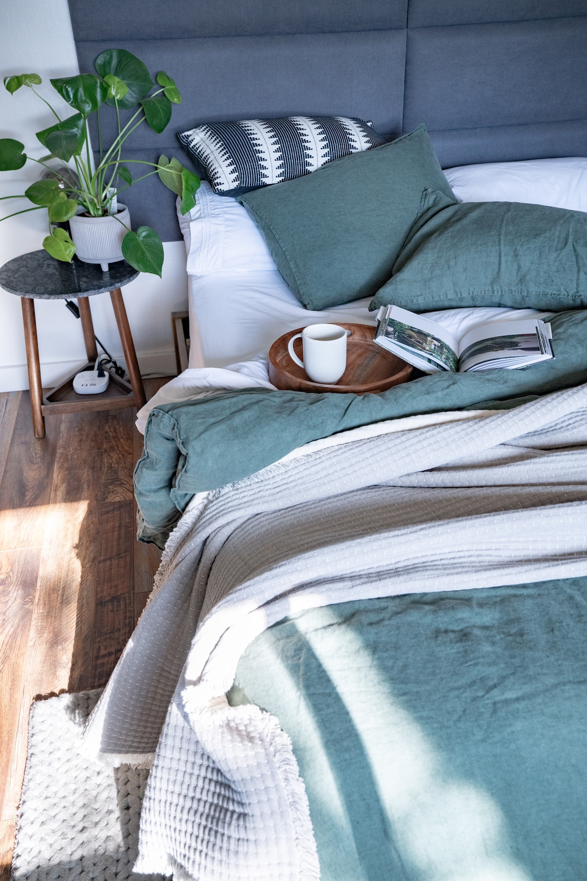 A cozy sleeping arrangement features a spacious bed layered with soft blankets in muted green tones. A plant adds a touch of nature, while a bedside table holds a book and a steaming mug. Natural light gently illuminates the inviting space.