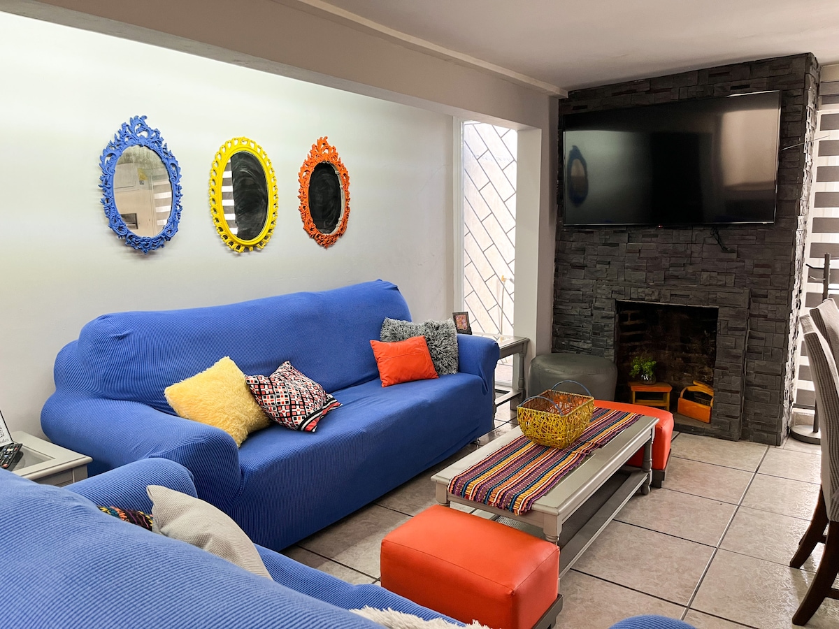 A spacious living area features a large, blue couch adorned with decorative pillows of various colors. Brightly colored mirrors hang on the wall, complemented by a coffee table with a colorful runner. A television is mounted above a stone fireplace, enhancing the cozy environment.