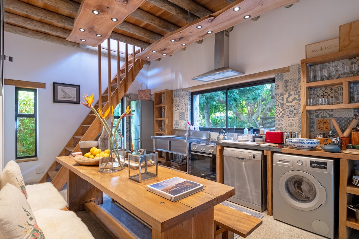 An open kitchen and dining area is shown, highlighting a large hardwood table surrounded by natural light. The kitchen is equipped with modern appliances, including a dishwasher and washing machine. Decorative tiles and natural elements create a cohesive design, alongside views of surrounding greenery.