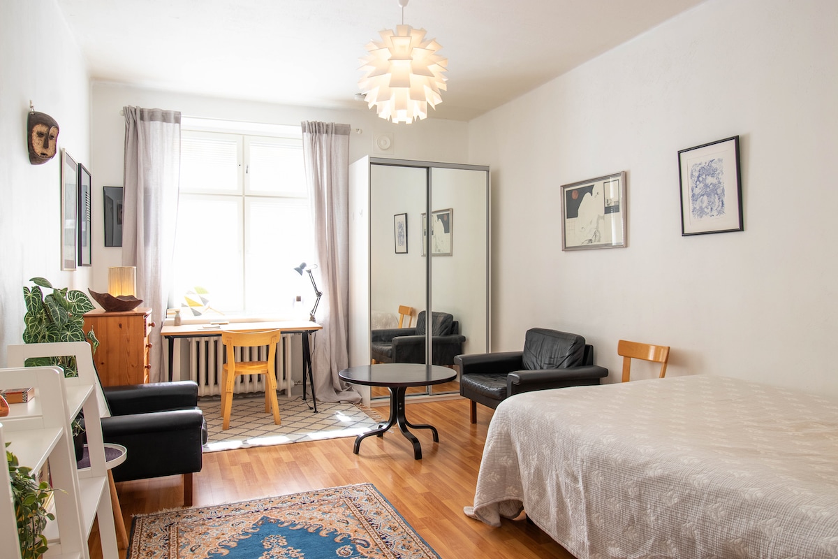A well-lit living space features a spacious bed dressed in light-colored bedding. A black sofa and a chair are situated around a circular table. A desk with a lamp is positioned near a large window, adorned with soft gray curtains. Artwork decorates the walls.
