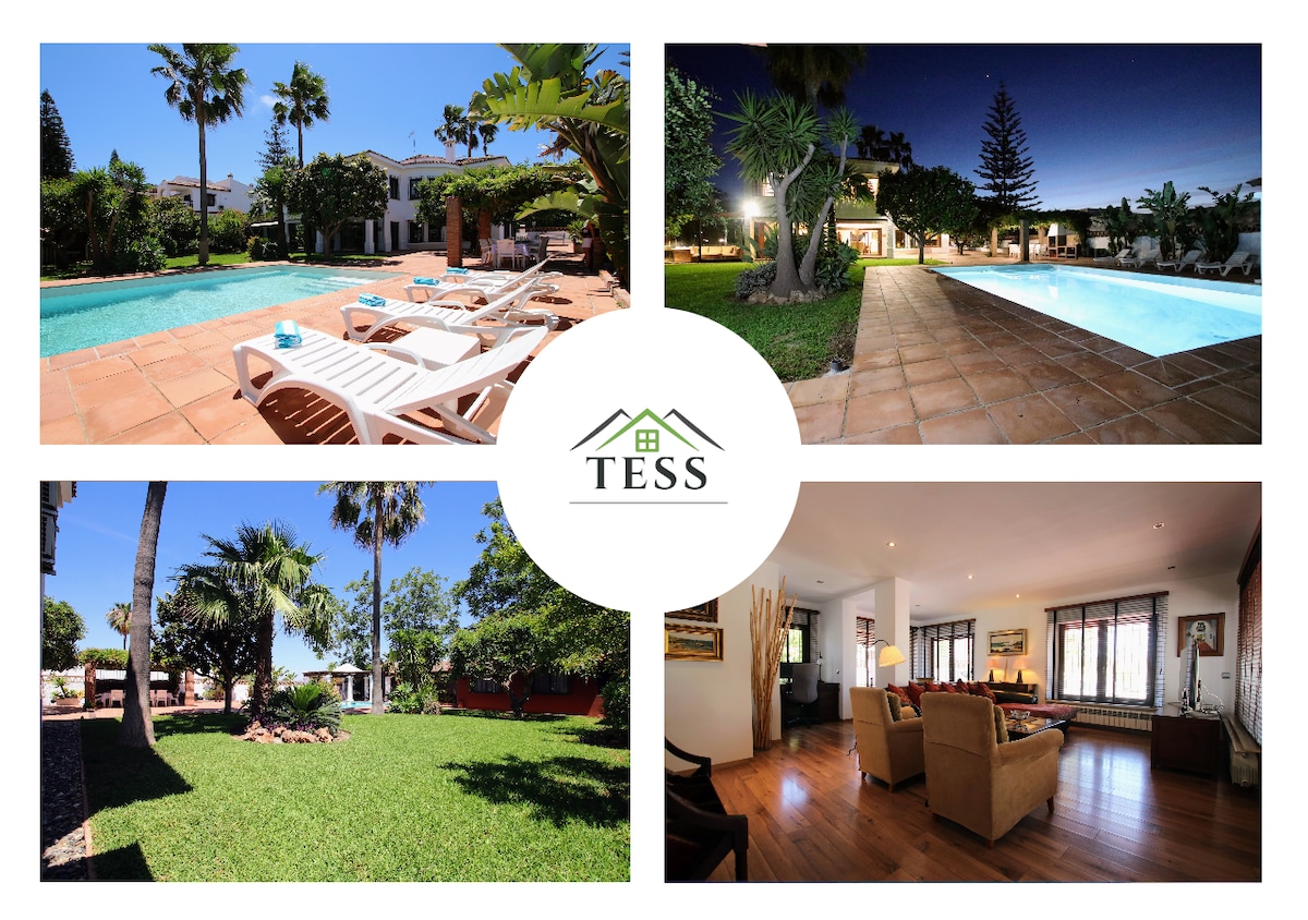 A spacious outdoor area features a large swimming pool surrounded by sunbeds and lush greenery. The garden is well-maintained, showcasing various plants and trees, creating a serene environment. The area is inviting for relaxation under the sun or in shaded spaces.