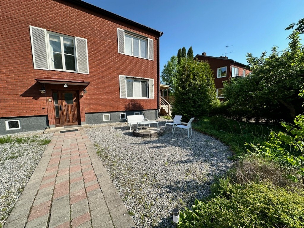 A well-maintained outdoor area features a gravel path leading to a seating arrangement of white chairs surrounding a fire pit. Lush greenery and small shrubs border the space, with a brick exterior of a two-story building visible in the background.