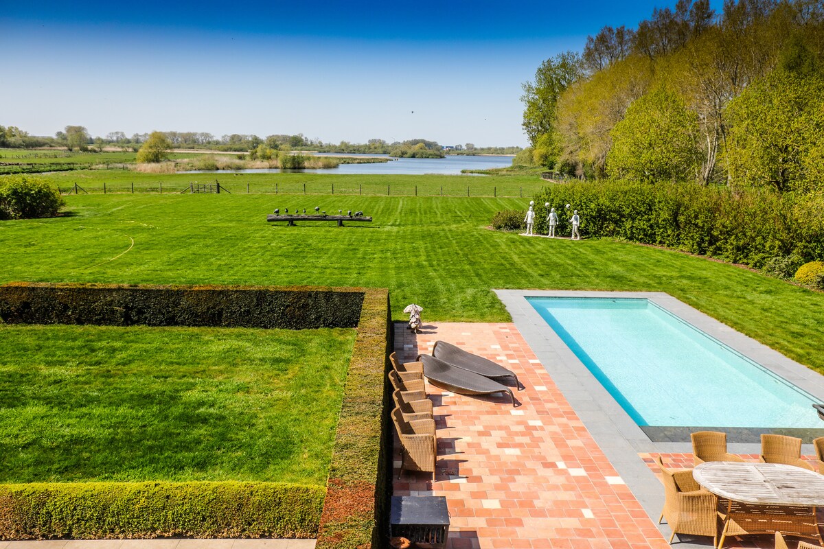 A serene view showcases a well-maintained lawn leading to a calm body of water. A rectangular pool features clear blue water and is bordered by a stone patio. A seating area with garden chairs and an outdoor dining table is situated beside the pool.