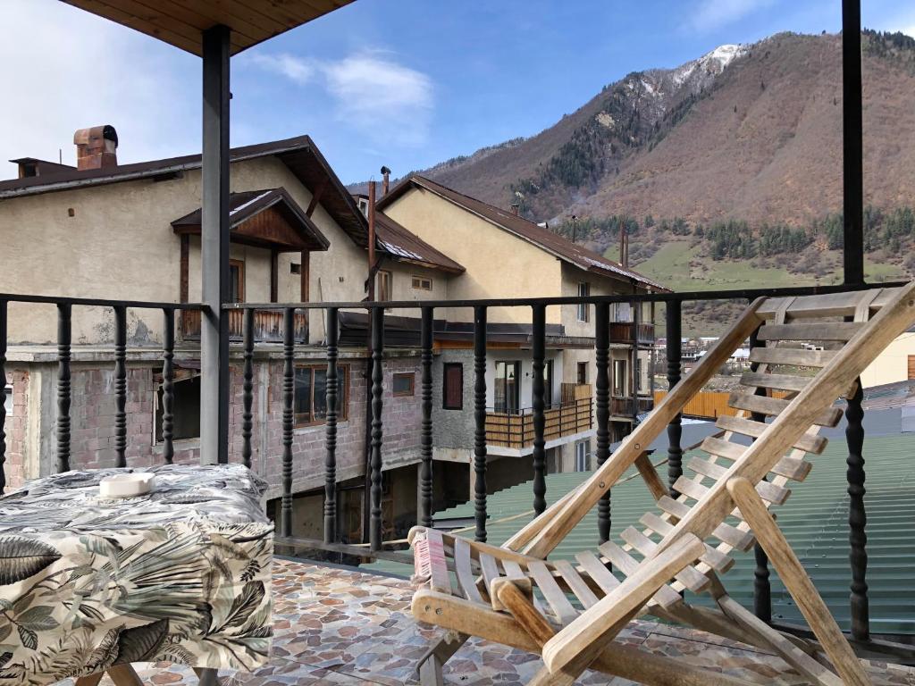 A balcony area is presented with a wooden lounge chair positioned for relaxation. A small table with a decorative cloth is visible. The backdrop showcases mountain scenery, with multiple buildings peeking through. A partially cloudy sky adds to the peaceful atmosphere.