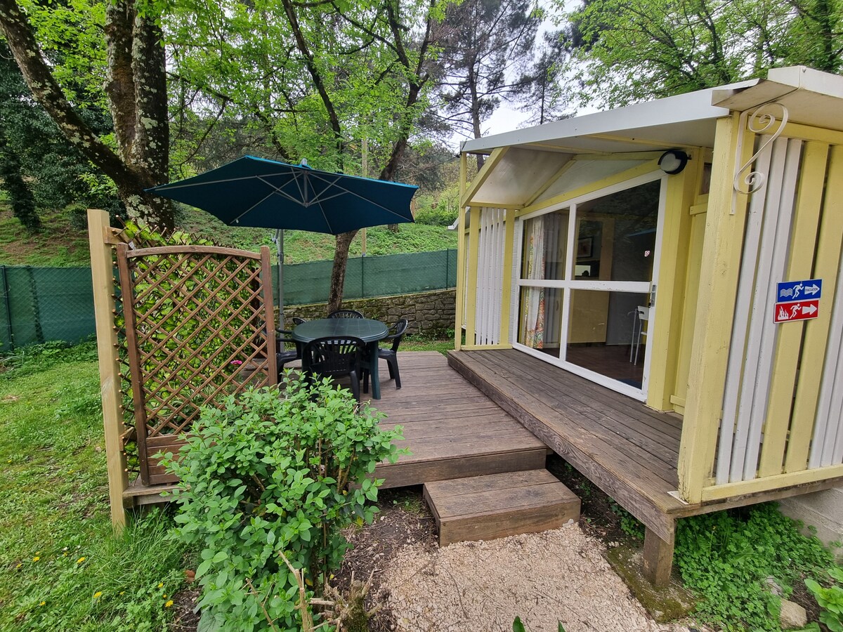 A shaded outdoor area is presented with a wooden deck, featuring a table and four black chairs beneath a large umbrella. Lush greenery surrounds the space, and a lattice structure adds texture to the setting.