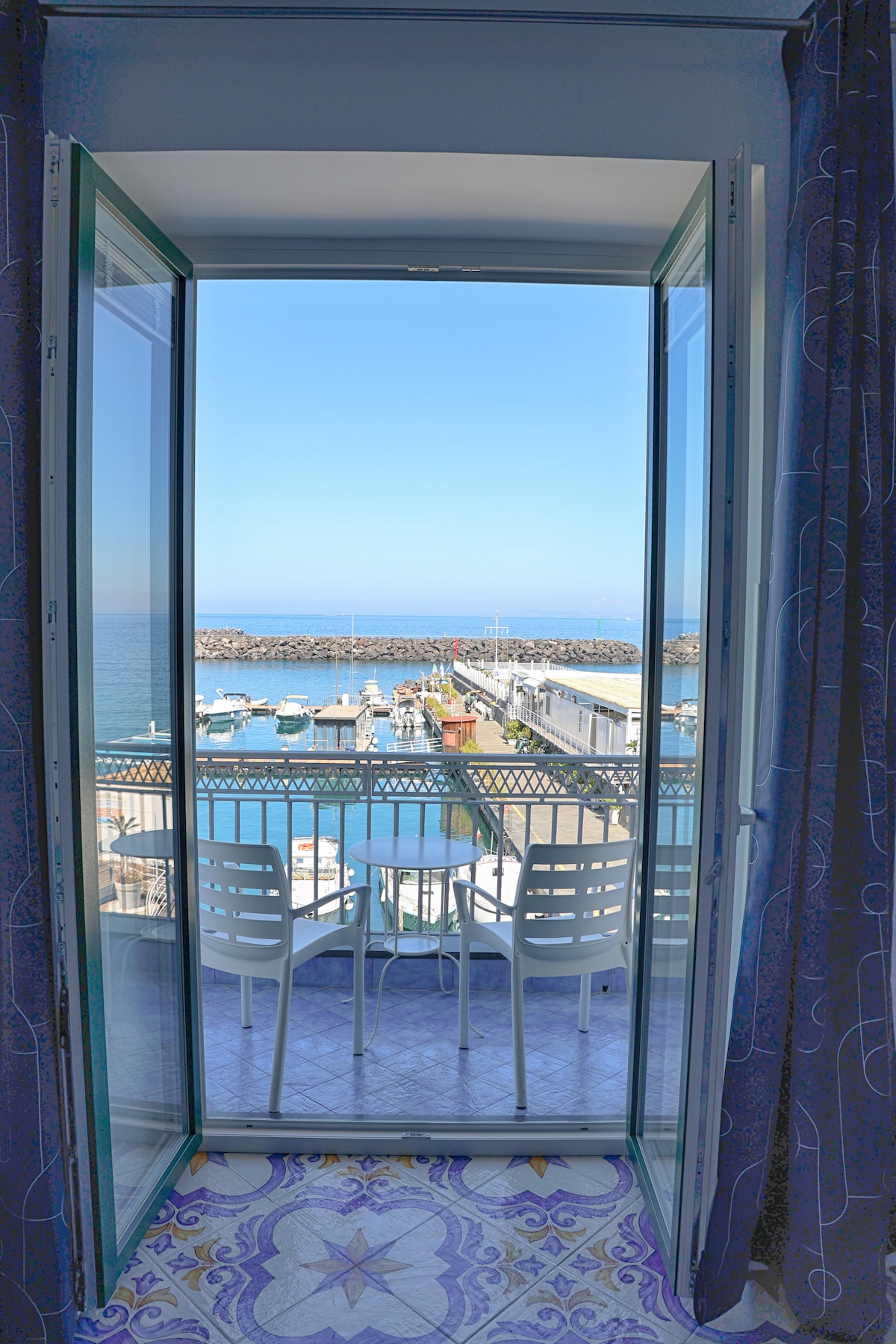 A balcony doorway provides a view of the marina, featuring numerous boats docked in the water. Two light-colored chairs are positioned around a small table on the balcony, with vibrant tiles visible on the floor. The scene is set against a clear blue sky.