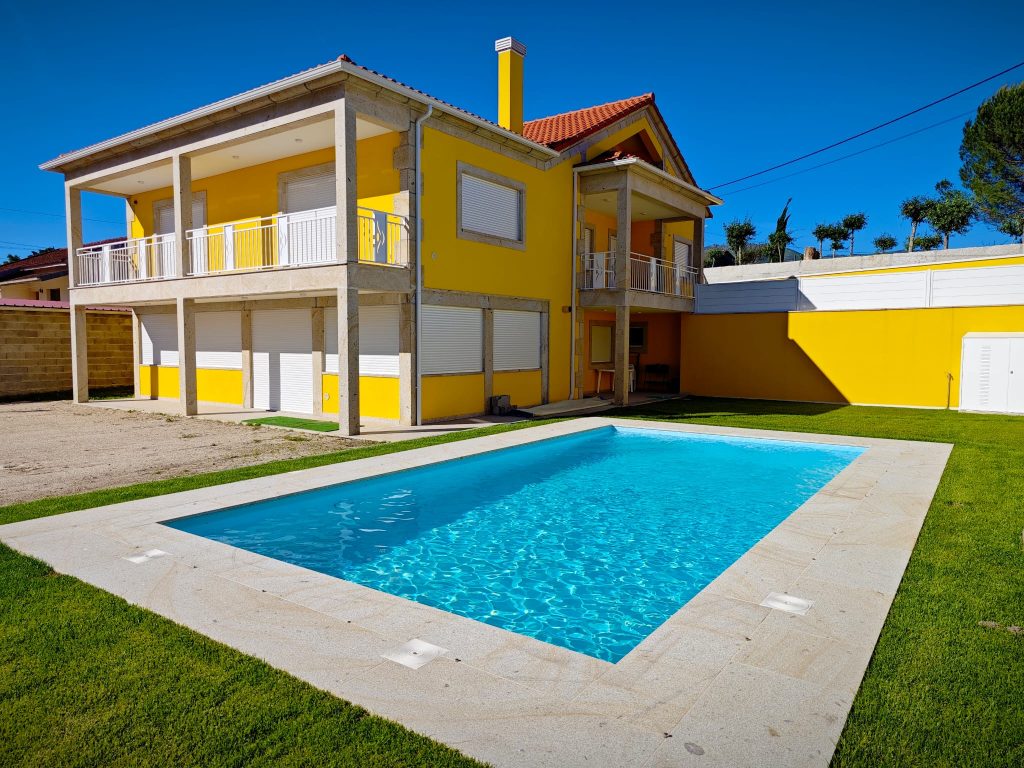 A bright yellow two-story house is complemented by a serene rectangular swimming pool. Lush green grass surrounds the pool area, providing a vibrant contrast. The property features balconies on both levels, framed by white railings, enhancing the overall modern aesthetic.